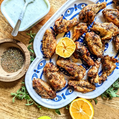 Mediterranean Chicken Wings served on a blue and white evil eye platter. The platter is surrounded by sprigs of fresh oregano and lemon wedges. A dish of feta and mint dip and a bowl of spice mix can be seen in the frame.