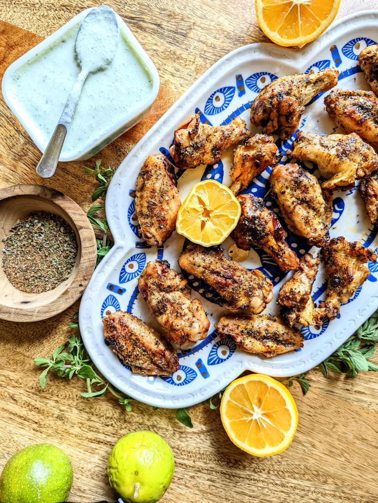 Mediterranean Chicken Wings served on a blue and white evil eye platter. The platter is surrounded by sprigs of fresh oregano and lemon wedges. A dish of feta and mint dip and a bowl of spice mix can be seen in the frame.