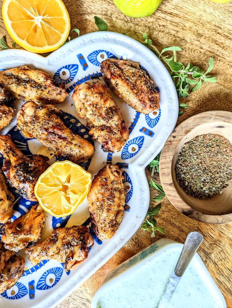 A closeup of Mediterranean Chicken Wings served on a blue and white evil eye platter. The platter is surrounded by sprigs of fresh oregano and lemon wedges. A small bowl of spice mix and a small bowl of dip can also be seen in the frame.