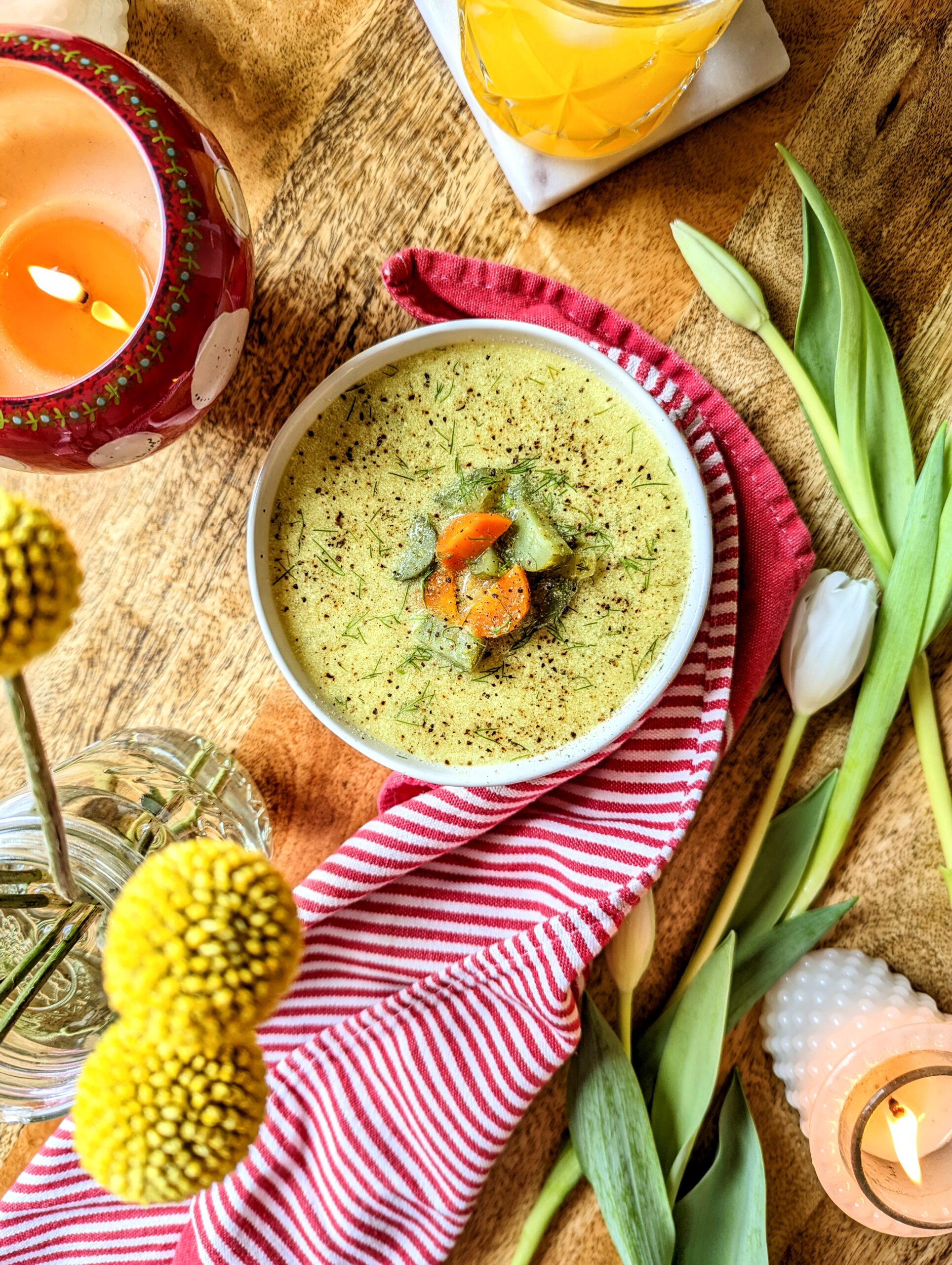 A bowl of soup with green dill pickles, orange carrots, and black pepper. White and yellow flowers, along with lite candles can be seen.