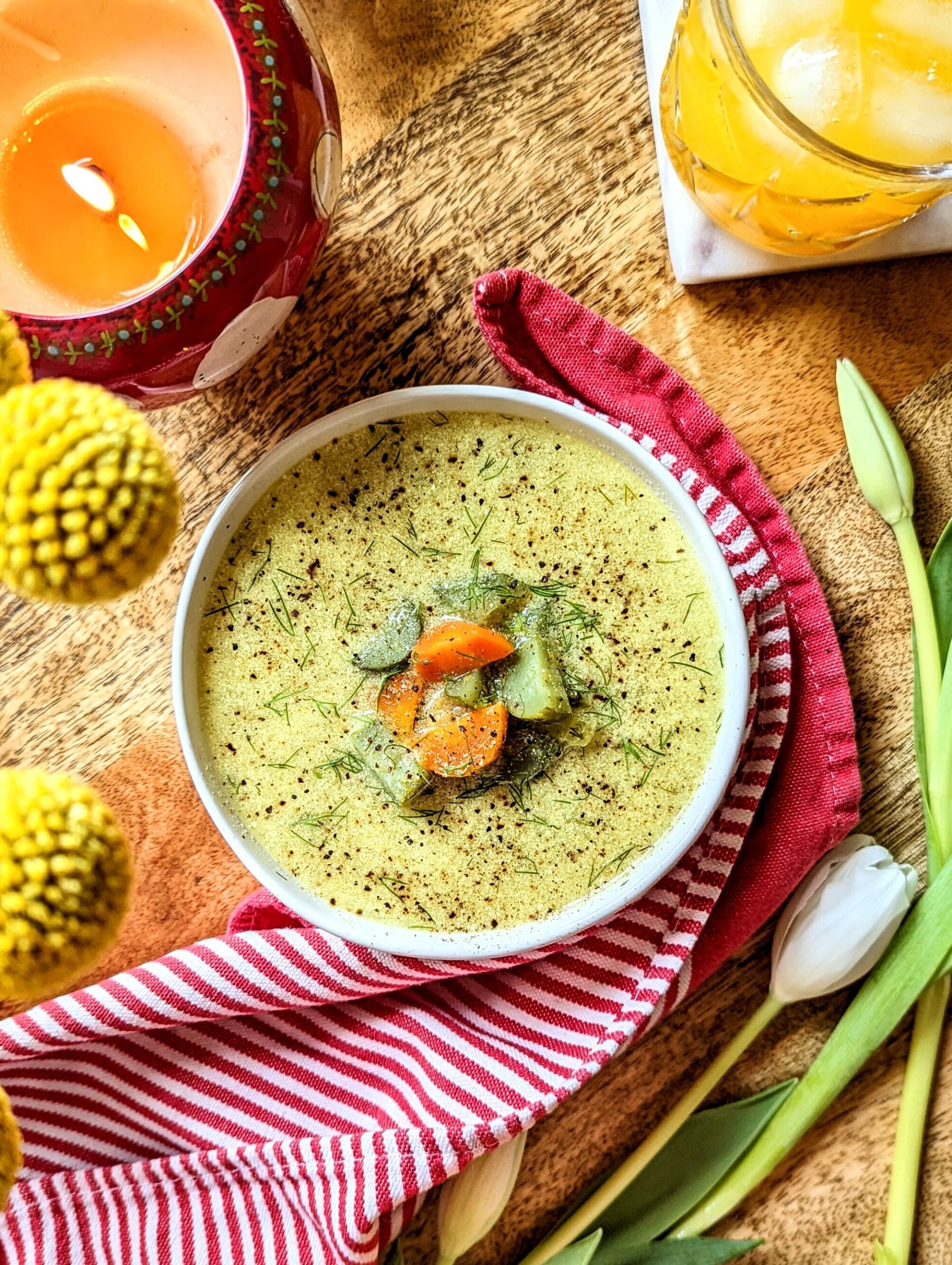 A birds eye view of a table set for lunch. A bowl of soup, a glass of orange juice, lit candles, and spring florals can be seen around the bowl of Lazy Dill Pickle Soup.