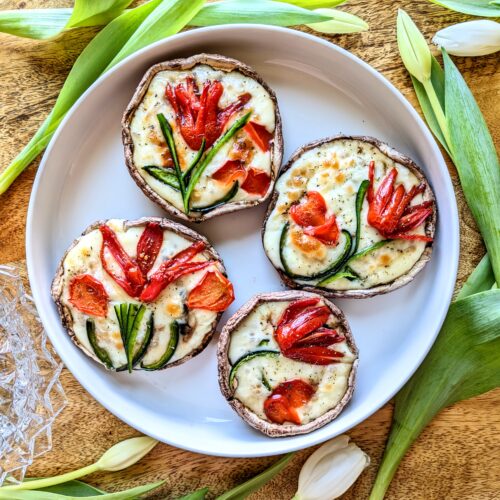 Four portobello mushrooms stuffed with mozzarella cheese, tomato, and bell pepper. The tomato and bell peppers are cut into shaped to look like tulips. Loose white tulips surround the large plate.