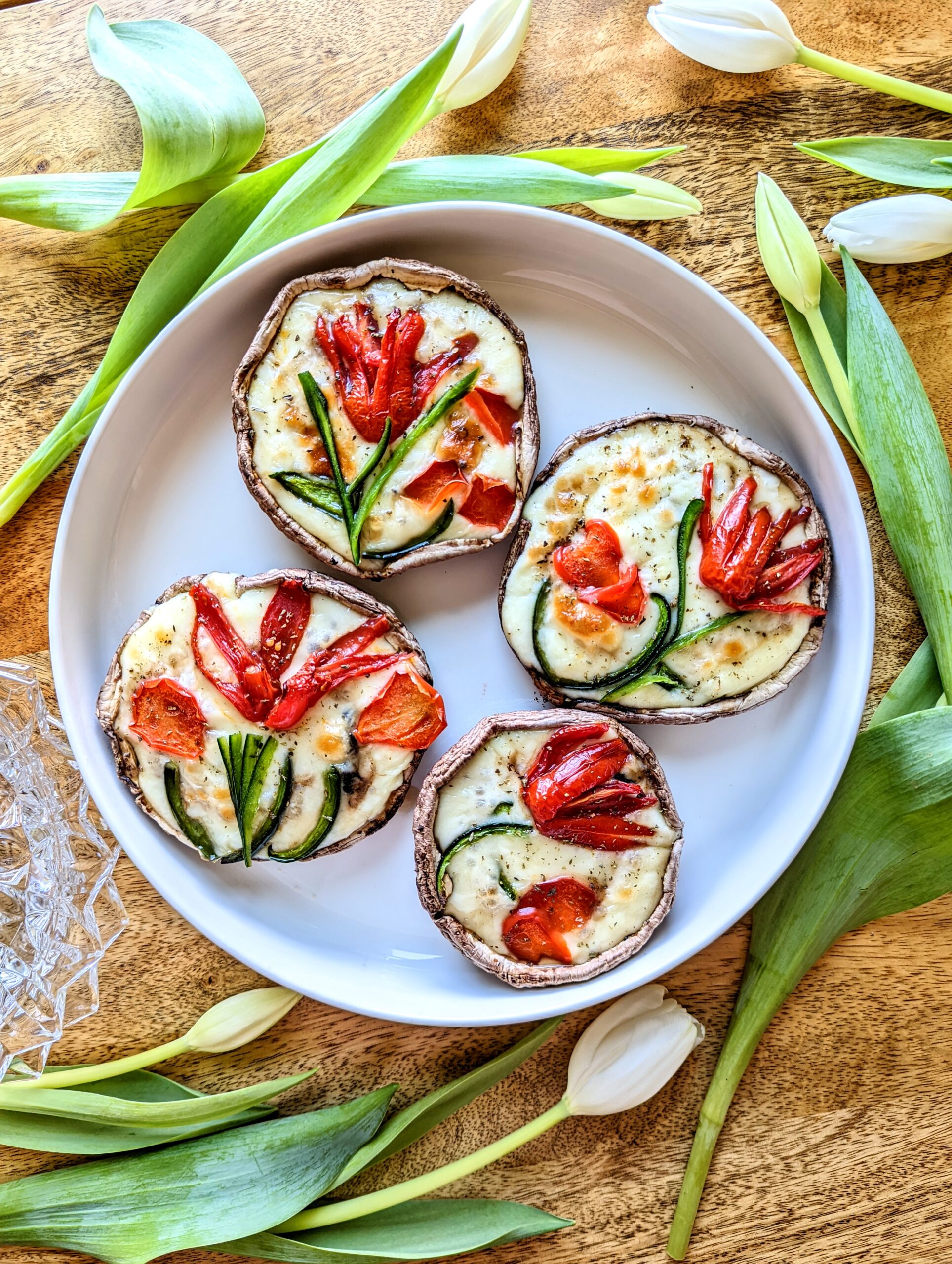 Four portobello mushrooms stuffed with mozzarella cheese, tomato, and bell pepper. The tomato and bell peppers are cut into shaped to look like tulips. Loose white tulips surround the large plate.