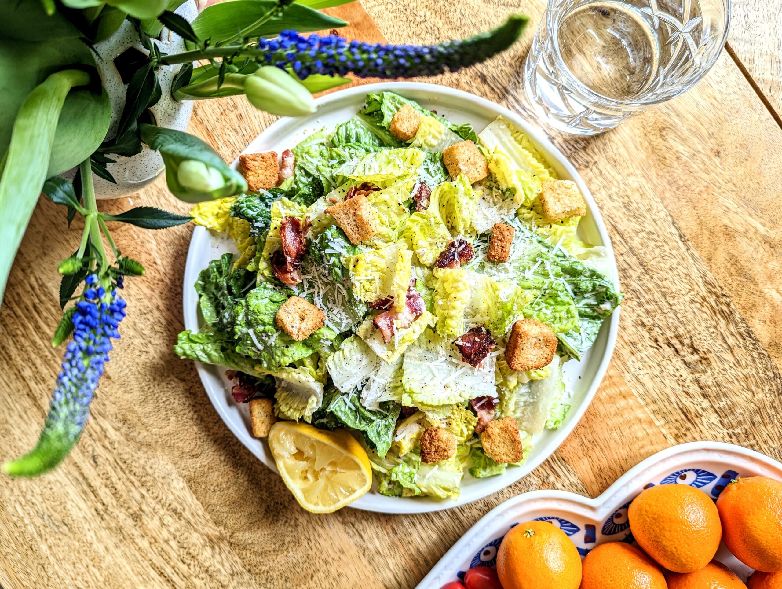 A large plate of Caesar Salad with bacon and homemade croutons. Purple Veronica flowers are seen in the top left corner and a platter of orange mandarins can be seen in the bottom right corner.
