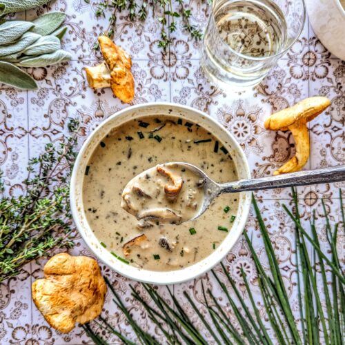 A spoonful of Wild Mushroom Soup, surround by fresh sage, thyme, chives, and whole chanterelle mushrooms. A short glass of white wine can be seen in the top right corner.