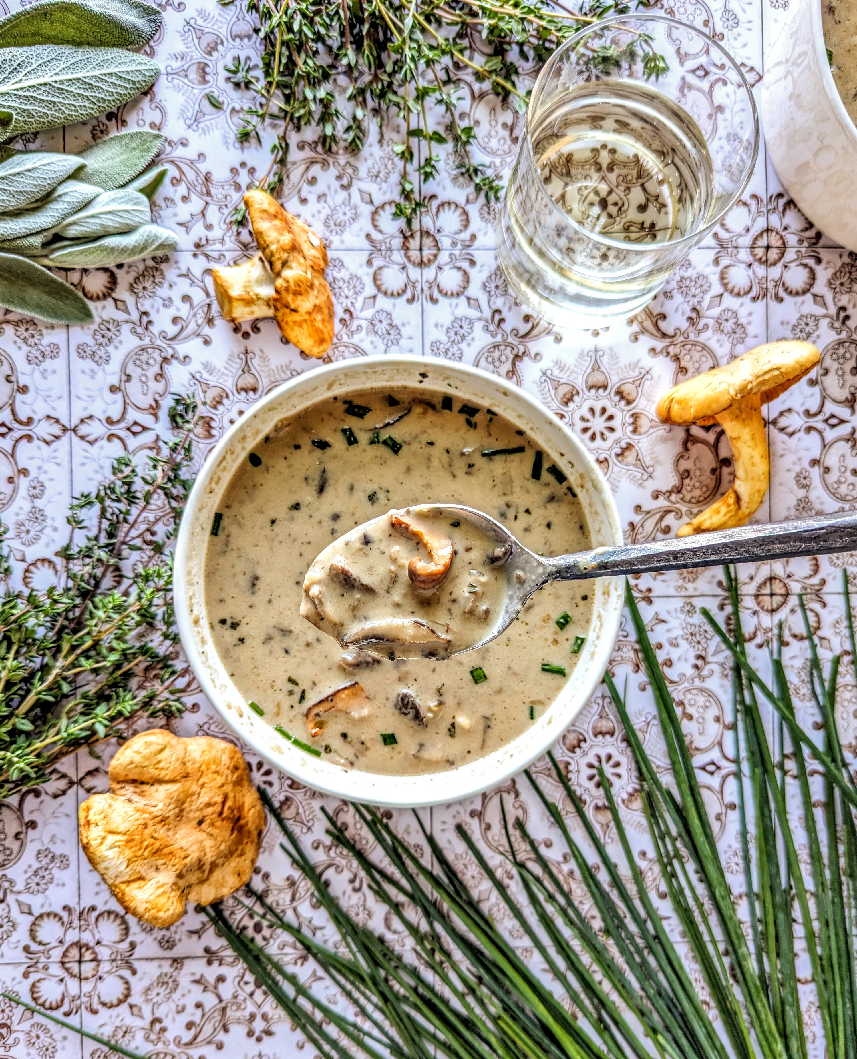 A spoonful of Wild Mushroom Soup, surround by fresh sage, thyme, chives, and whole chanterelle mushrooms. A short glass of white wine can be seen in the top right corner.