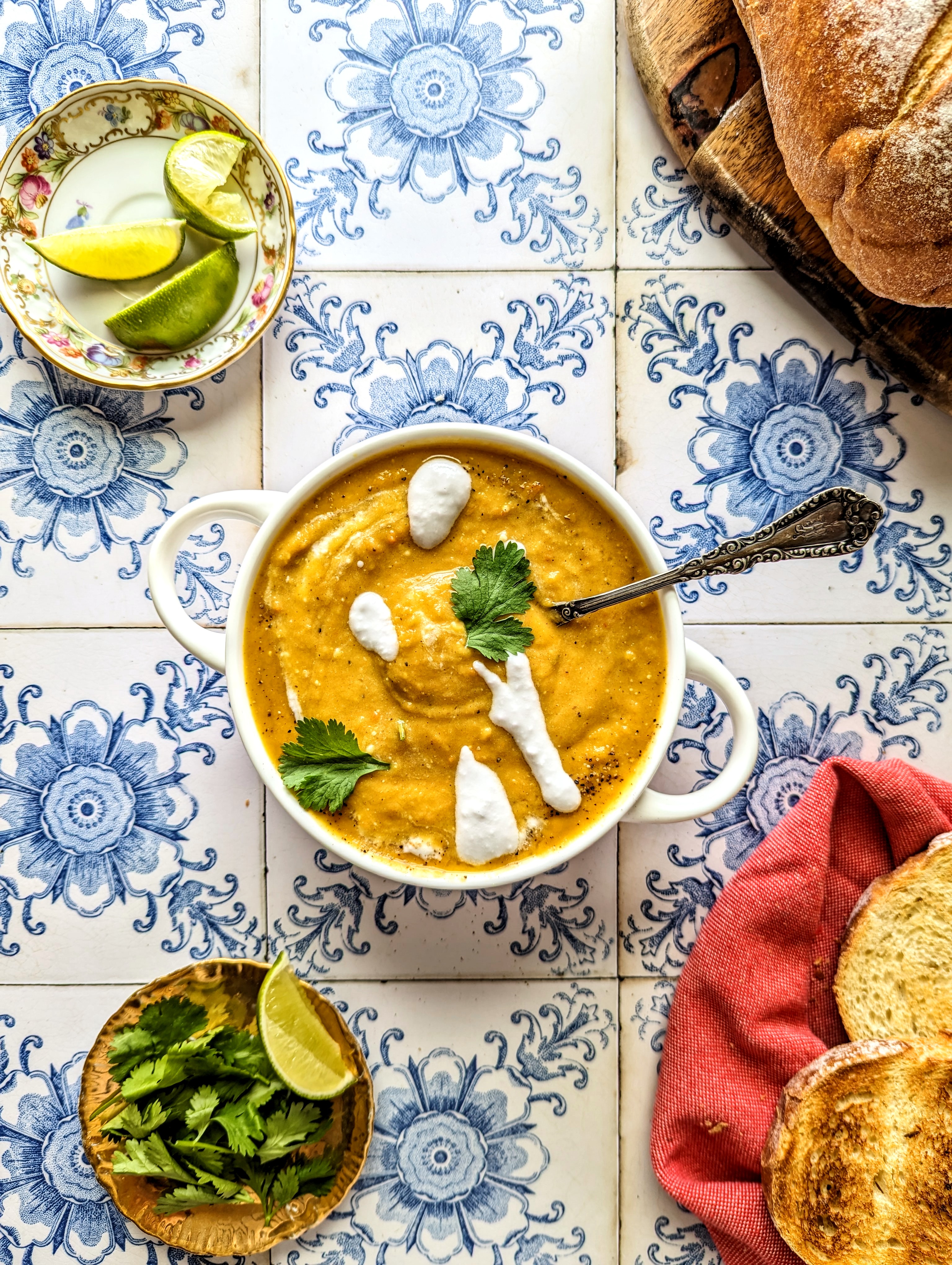 A spoon rests in a bowl of vibrant orange roasted squash and root vegetable soup. The soup is topped with white coconut cream and green cilantro.