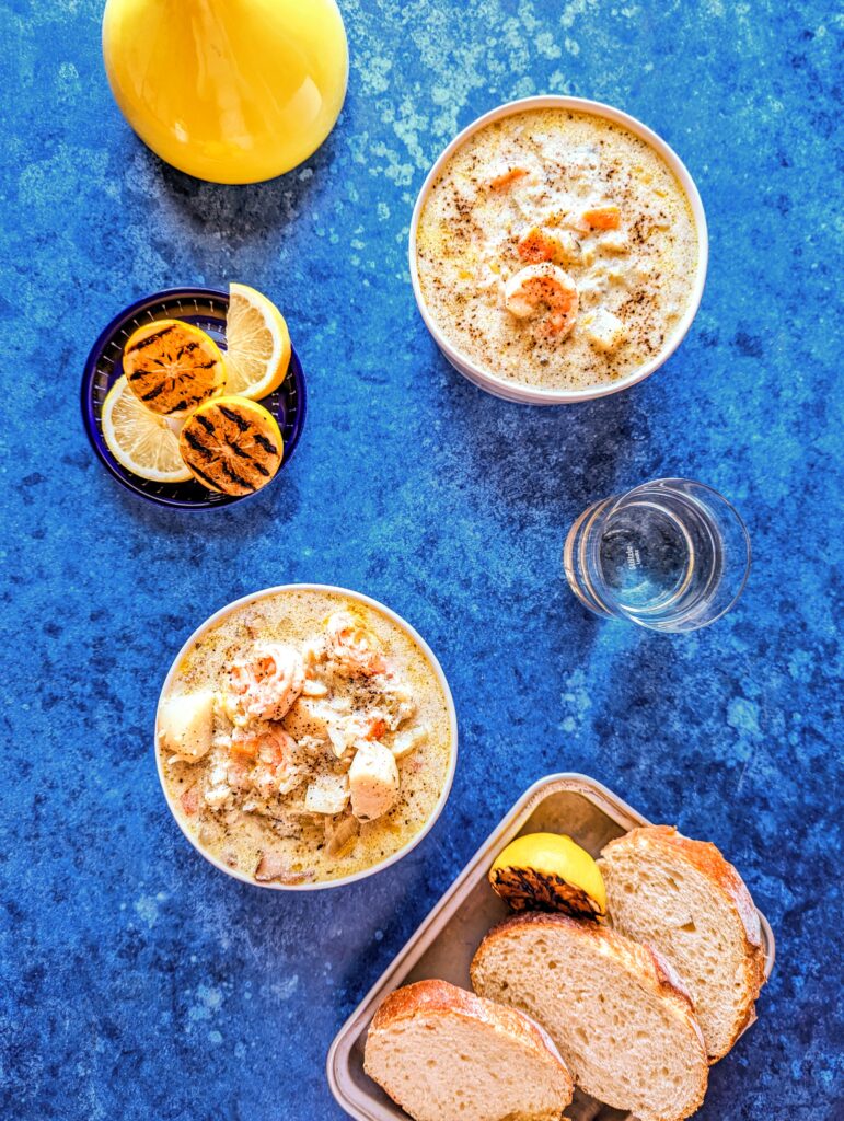 A flat lay of two bowl of Nova Scotia Seafood Chowder with a vibrant blue backdrop. A plate of grilled lemons and another of toasted bread are featured in the shot.