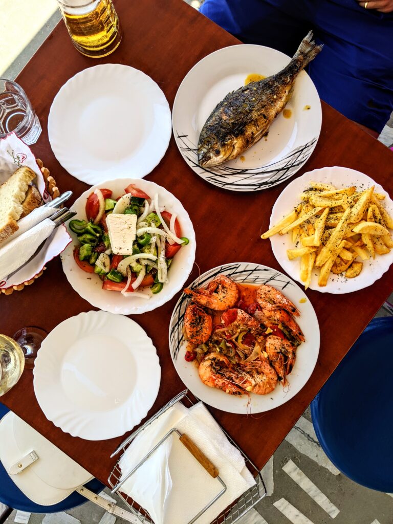A table full of Greek salad, fresh french fries, whole grilled fish, and grilled shrimp.