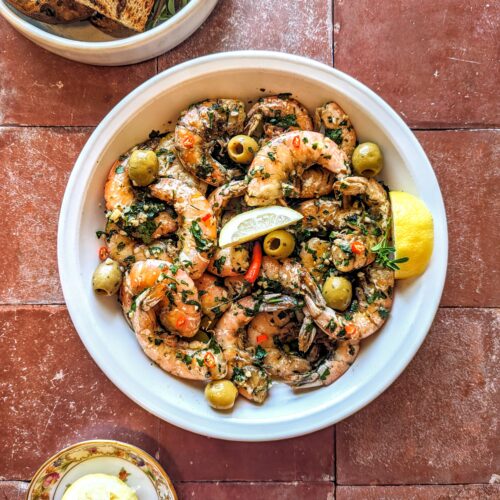 A large bowl of Grilled Garlic and Chili Shrimp with green parsley, green olived, fresh lemon wedges, and grilled sourdough toasts.