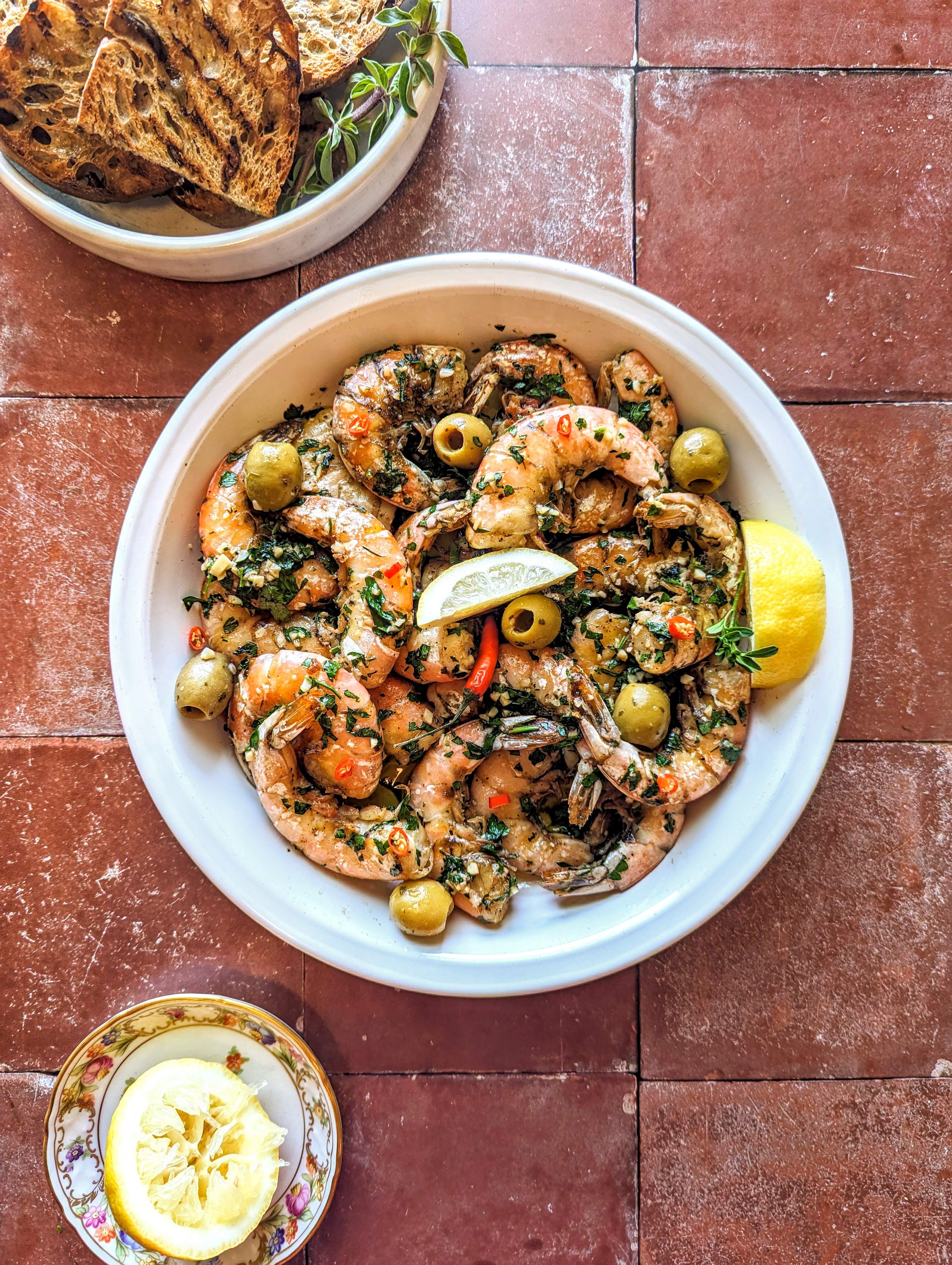 A large bowl of Grilled Garlic and Chili Shrimp with green parsley, green olived, fresh lemon wedges, and grilled sourdough toasts.