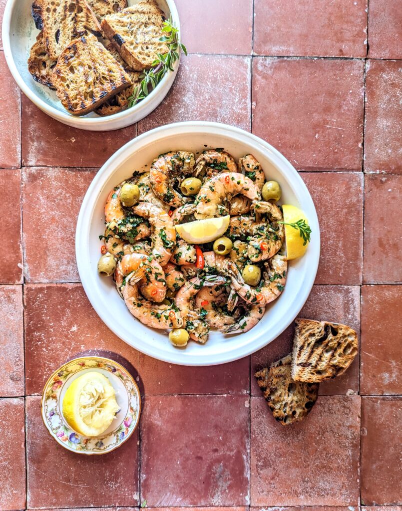 A terracota table with a big bowl of Garlic Chili Shrimp and Sourdough Toasts.