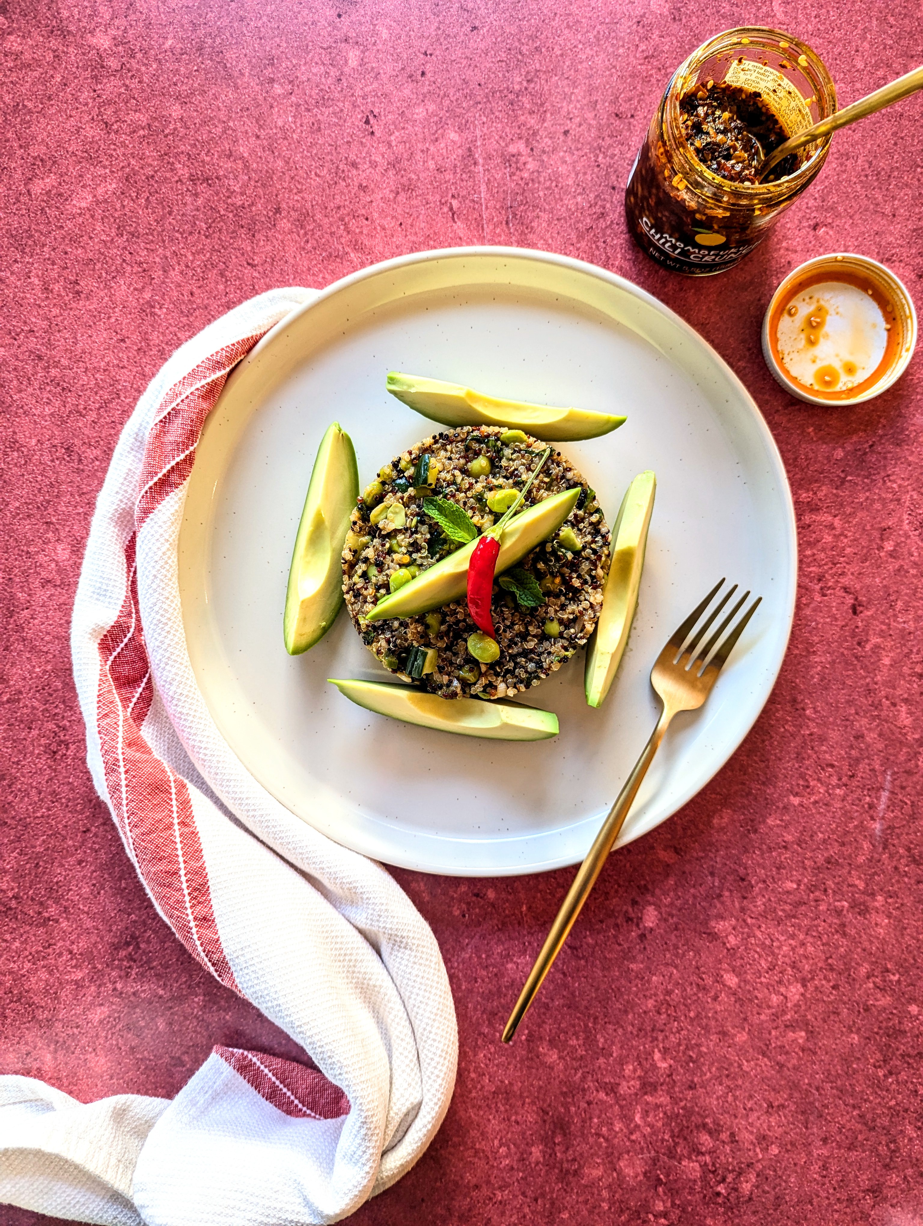 A flat-lay with a red background and white plate of Spicy Quinoa Salad. An opened jar of Momofuku chili crunch and a red & white dish towel are visible.