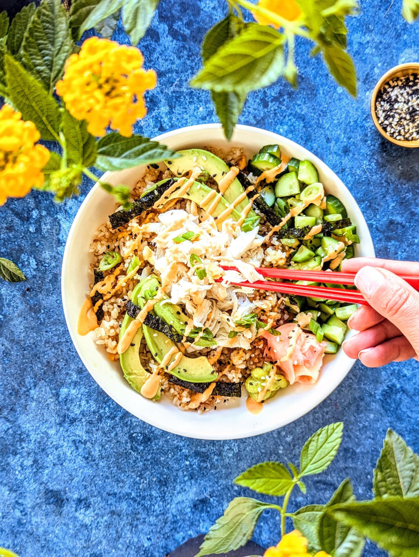 Red chopsticks digging into a Crispy Californian Roll Bowl. A small gold bowl of white and black sesame seeds, yellow flowers, and greenery, pop on a bright blue background.